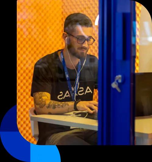 Colaborador do Asaas utiliza uma das cabines de reunião da sede. Ele está usando fones de ouvido enquanto mexe no computador.