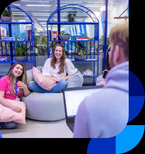 Colaboradores do Asaas conversam na área criativa. Há duas mulheres sorrindo, uma está sentada em um puff e a outra de joelhos no chão, com uma almofada no colo. No canto inferior direito, há um homem de costas, em desfoque. Ao fundo, estão duas estantes coloridas em azul e algumas plantas.